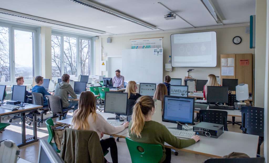 Schülerinnen und Schüler sitzen vor Monitoren im Klassenzimmer. Der Lehrer sitzt am Lehrertisch vor einem Monitor.