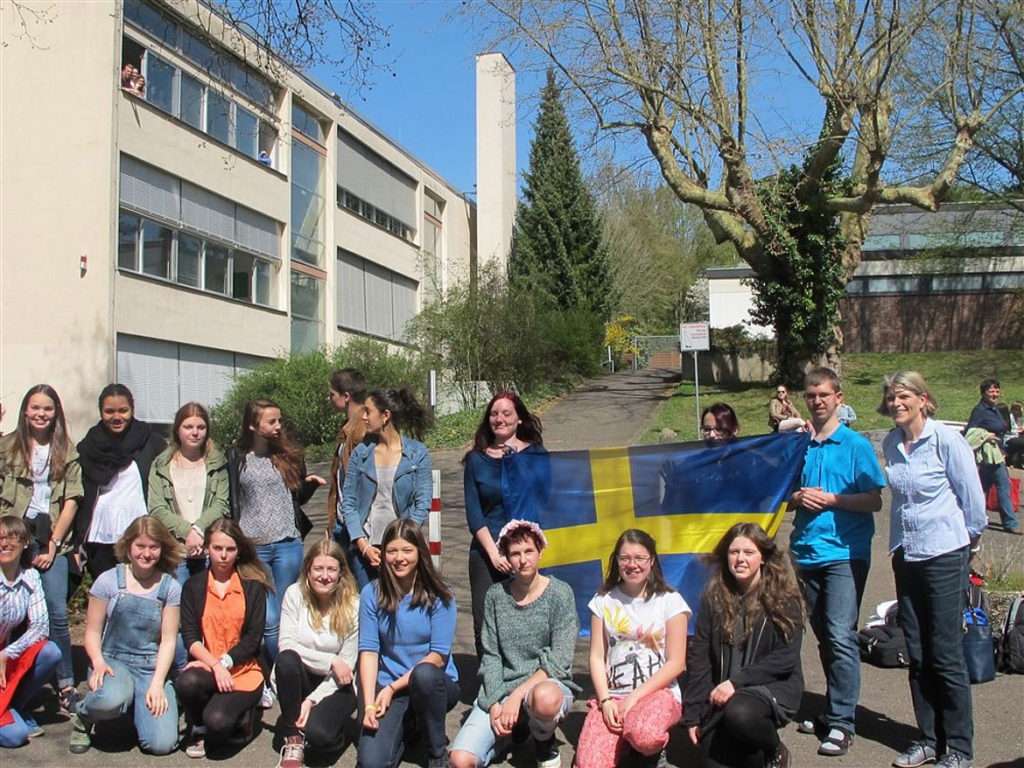 Eine Lehrerin, Schülerinnen und Schüler der Schwedisch AG stehen in einer großen Gruppe zusammen. Zwei Schüler halten gemeinsam die schwedische Flagge nach oben.